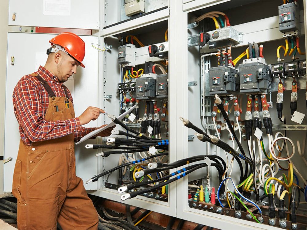 Elektrikář v oranžové přilbě kontroluje zapojení kabelů v rozvaděči průmyslové automatizace.